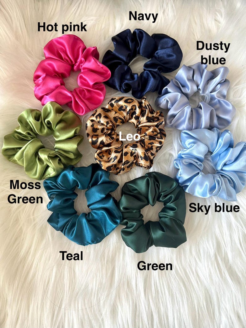 May include: A collection of satin scrunchies in various colors, including hot pink, navy, dusty blue, moss green, teal, green, and sky blue. One scrunchie features a leopard print. The scrunchies are displayed on a white, fluffy surface.
