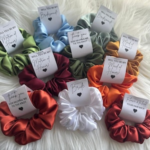 May include: A collection of satin scrunchies in various colors, including green, blue, orange, and red. Each scrunchie has a small white tag with text such as "Will you be my Bridesmaid?" and "Bride" with a heart symbol. The scrunchies are arranged on a white, fluffy surface.