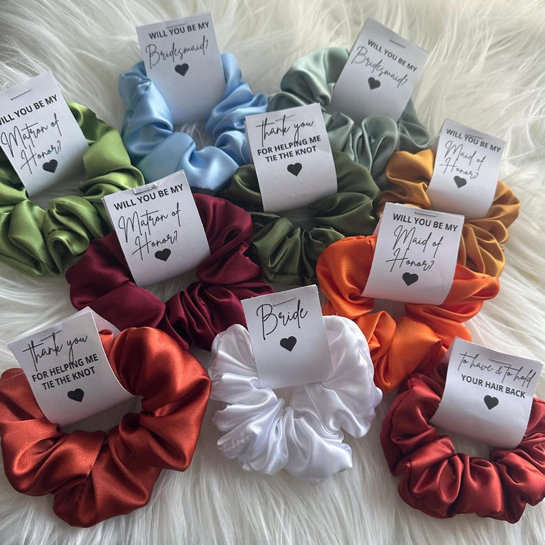 May include: A collection of satin scrunchies in various colors, including green, blue, orange, and red. Each scrunchie has a small white tag with text such as "Will you be my Bridesmaid?" and "Bride" with a heart symbol. The scrunchies are arranged on a white, fluffy surface.