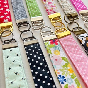 May include: Assortment of colorful fabric keychains with different patterns, including polka dots, floral prints, and geometric designs. Each keychain features a metal key ring.