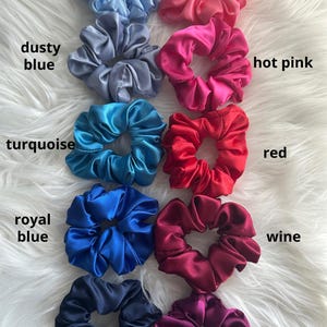 May include: A collection of satin scrunchies in various colors, including sky blue, dusty blue, turquoise, royal blue, navy, coral rose, hot pink, red, wine, and magenta. Each scrunchie is labeled with its corresponding color name.