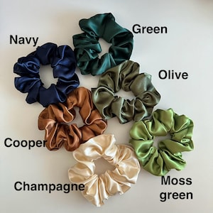 May include: A collection of satin scrunchies in various colors, including navy, green, olive, copper, champagne, and moss green. Each scrunchie is labeled with its respective color name. These hair accessories are designed to hold hair in place.