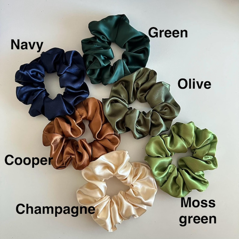 May include: A collection of satin scrunchies in various colors, including navy, green, olive, copper, champagne, and moss green. Each scrunchie is labeled with its respective color name. These hair accessories are designed to hold hair in place.