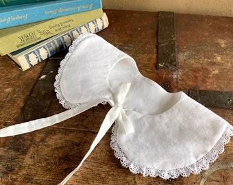 May include: White cotton collar with lace trim and a white satin ribbon tie. The collar is lying on a wooden surface.