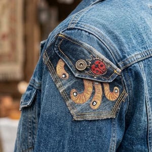 Könnte beinhalten: Eine mittelblaue Jeansjacke mit einem dekorativen Aufnäher am Oberarm. Der Aufnäher zeigt einen roten Marienkäfer und goldfarbene, stilisierte Designs. Die Jacke hat eine geknöpfte Tasche und eine geknöpfte Manschette.