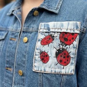 Puede incluir: Chaqueta vaquera azul claro con un bolsillo con un diseño pintado a mano de mariquitas rojas y negras. La chaqueta tiene botones dorados y un cuello clásico.