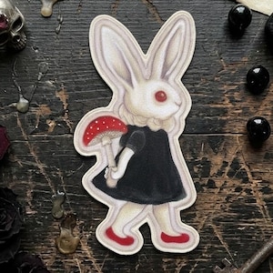 Può includere: Un adesivo fustellato raffigurante un coniglio bianco con occhi rossi, che indossa un vestito nero e scarpe rosse, mentre tiene in mano un fungo rosso a pois bianchi. L'illustrazione ha un contorno chiaro ed è una grafica adesiva piatta.