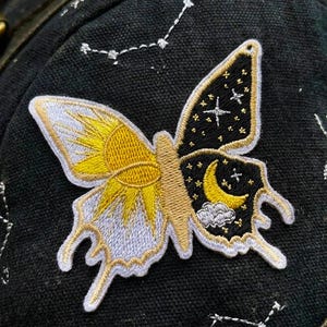May include: An embroidered butterfly patch on a black fabric garment. The left wing features a yellow sun design, while the right wing displays a black night sky with a yellow crescent moon, stars, and a small cloud. White constellations are stitched nearby.