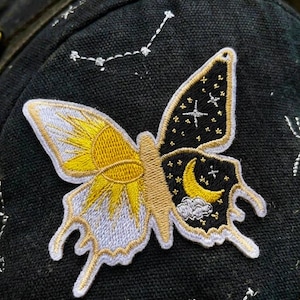 May include: Embroidered butterfly patch with a celestial design. One wing features a yellow sun, the other a night sky with stars and a crescent moon. The patch is sewn onto a black fabric, with embroidered constellations.