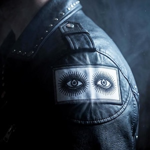 May include: Black leather jacket with silver studs and a patch featuring two stylized eyes. The jacket has a classic biker style with a notched collar and a zipper closure. The patch is rectangular and sewn onto the upper left sleeve.