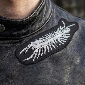Op de afbeelding: Zwarte leren jas met een duizendpoot patch. De patch is zwart met een wit duizendpoot ontwerp. De jas heeft een klassieke biker stijl met een reverskraag en zilveren knopen.
