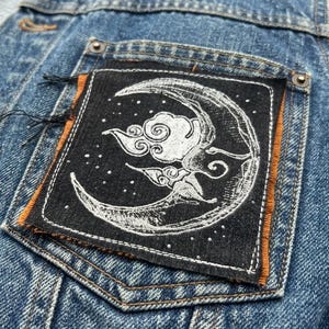 Puede incluir: Primer plano de una chaqueta vaquera azul con un parche decorativo. El parche es negro con un diseño de luna creciente, nube y estrellas en blanco. El parche tiene un borde naranja y está cosido en el bolsillo de la chaqueta.