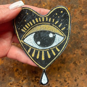 May include: Heart-shaped patch with an eye design. The patch features a black background with gold and white accents. The eye is white with a black pupil, and the design includes a gold border and a teardrop shape at the bottom.
