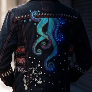 May include: A black denim jacket featuring a large teal octopus tentacle graphic on the back, accented with silver metal cone studs, multiple safety pins with small charms, and red stitched text patches that read "RIOT" and "NO FUTURE".