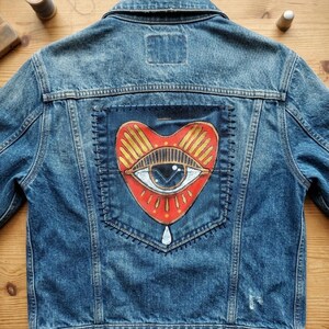May include: A blue denim jacket featuring a large, rectangular dark fabric patch sewn onto the back. The patch displays a hand-painted red heart shape containing a central eye motif with gold rays, black outlines, and a small white teardrop below.