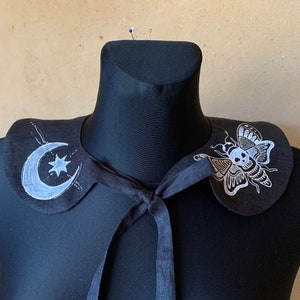 K&ouml;nnte beinhalten: Schwarzer Stoffkragen mit einem wei&szlig;en Halbmond- und Sternmuster auf der einen Seite und einem wei&szlig;en Schmetterling mit einem Totenkopfdesign auf der anderen Seite.