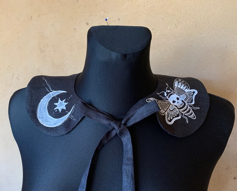 K&ouml;nnte beinhalten: Schwarzer Stoffkragen mit einem wei&szlig;en Halbmond- und Sternmuster auf der einen Seite und einem wei&szlig;en Schmetterling mit einem Totenkopfdesign auf der anderen Seite.