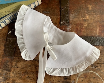 May include: A white linen collar with a ruffled edge and a white satin ribbon tie. The collar is lying on a wooden surface.