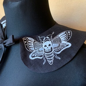 K&ouml;nnte beinhalten: Schwarzer Stoffkragen mit einem wei&szlig;en gestickten Totenkopf- und Mottenmotiv.