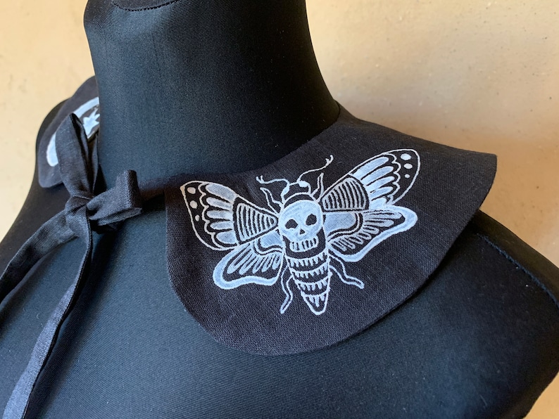 K&ouml;nnte beinhalten: Schwarzer Stoffkragen mit einem wei&szlig;en gestickten Totenkopf- und Mottenmotiv.