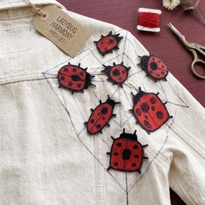 May include: A cream denim jacket with several red and black ladybug patches. The patches vary in size and design. A tag attached to the jacket reads "LADYBUG HARMONY - PATCH SET".
