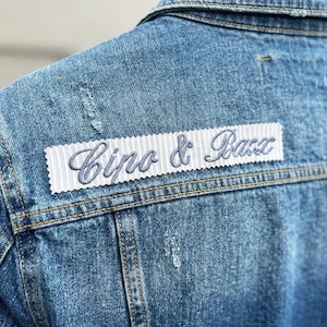 May include: Close-up of a light blue denim jacket with a distressed design. A white patch with the embroidered text "Cipo & Baxx" is sewn on the back of the jacket. The jacket has visible stitching and a classic collar.