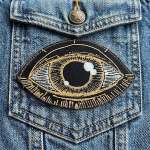 May include: A black and gold embroidered eye patch is sewn onto a denim jacket pocket. The patch features a detailed eye design with white and gold accents. The jacket has a star-shaped button.