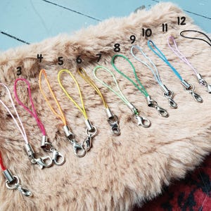 May include: A collection of twelve colorful wrist straps with silver-toned clasps. The straps are in various colors, including red, white, pink, orange, yellow, green, blue, and black. They are arranged on a fluffy, light brown surface.