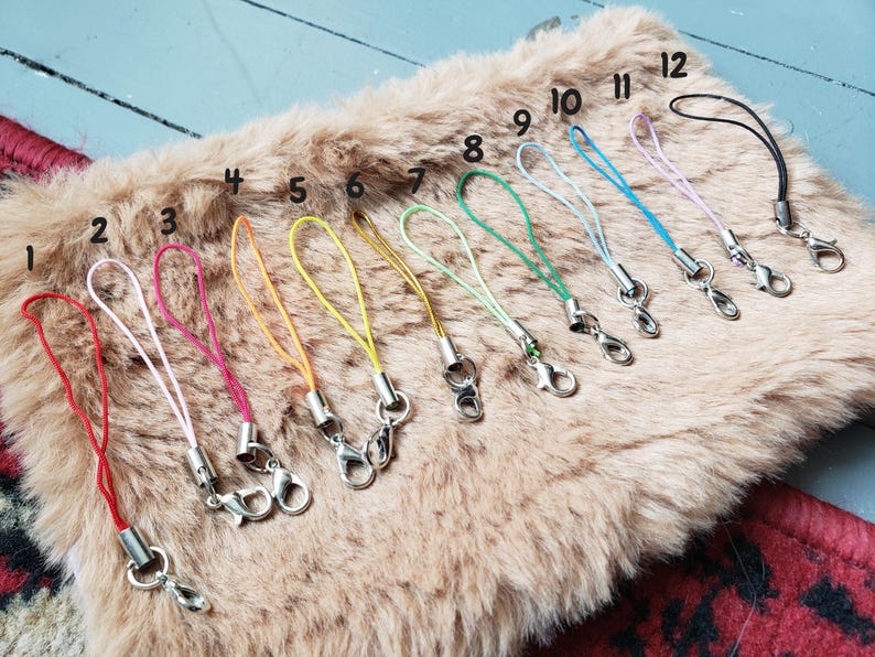May include: A collection of twelve colorful wrist straps with silver-toned clasps. The straps are in various colors, including red, white, pink, orange, yellow, green, blue, and black. They are arranged on a fluffy, light brown surface.