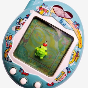 May include: A vintage Tamagotchi Town digital pet game in a light blue casing with colorful cartoon graphics. The screen displays a green background with a small green character. The device has white buttons and a heart-shaped opening.