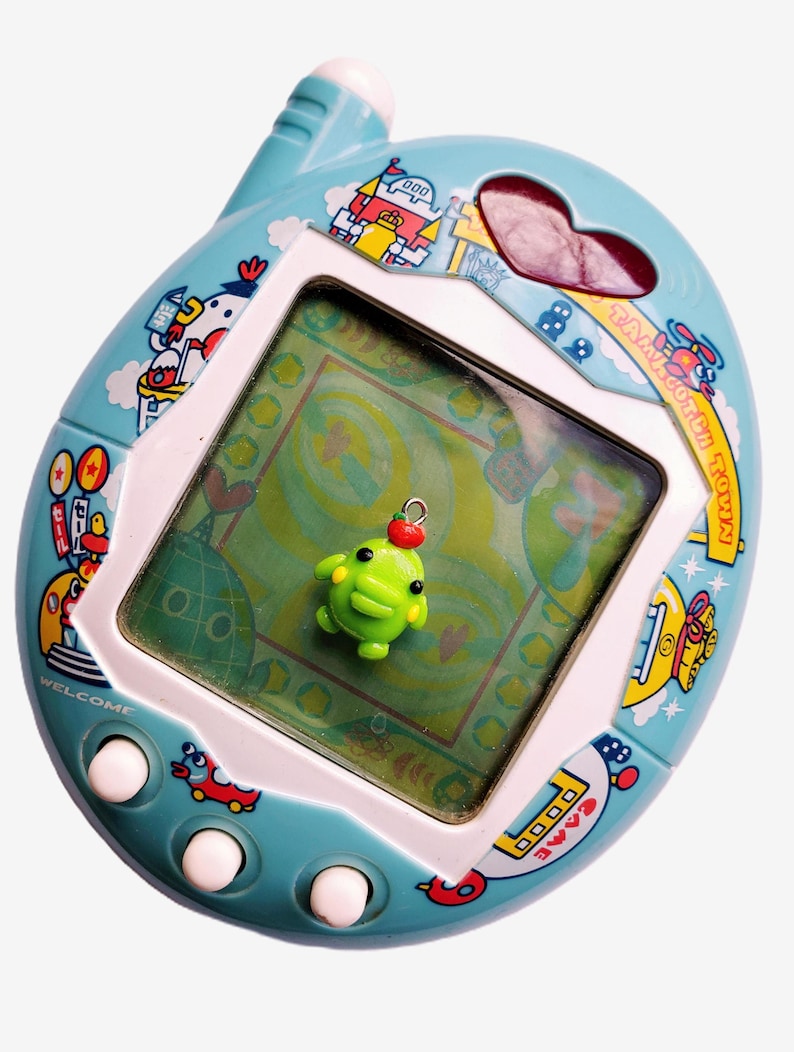 May include: A vintage Tamagotchi Town digital pet game in a light blue casing with colorful cartoon graphics. The screen displays a green background with a small green character. The device has white buttons and a heart-shaped opening.