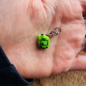May include: A small, green, handheld video game charm with a silver chain. The charm has a screen, black buttons, and pink cheeks. The charm is a miniature version of a retro video game.
