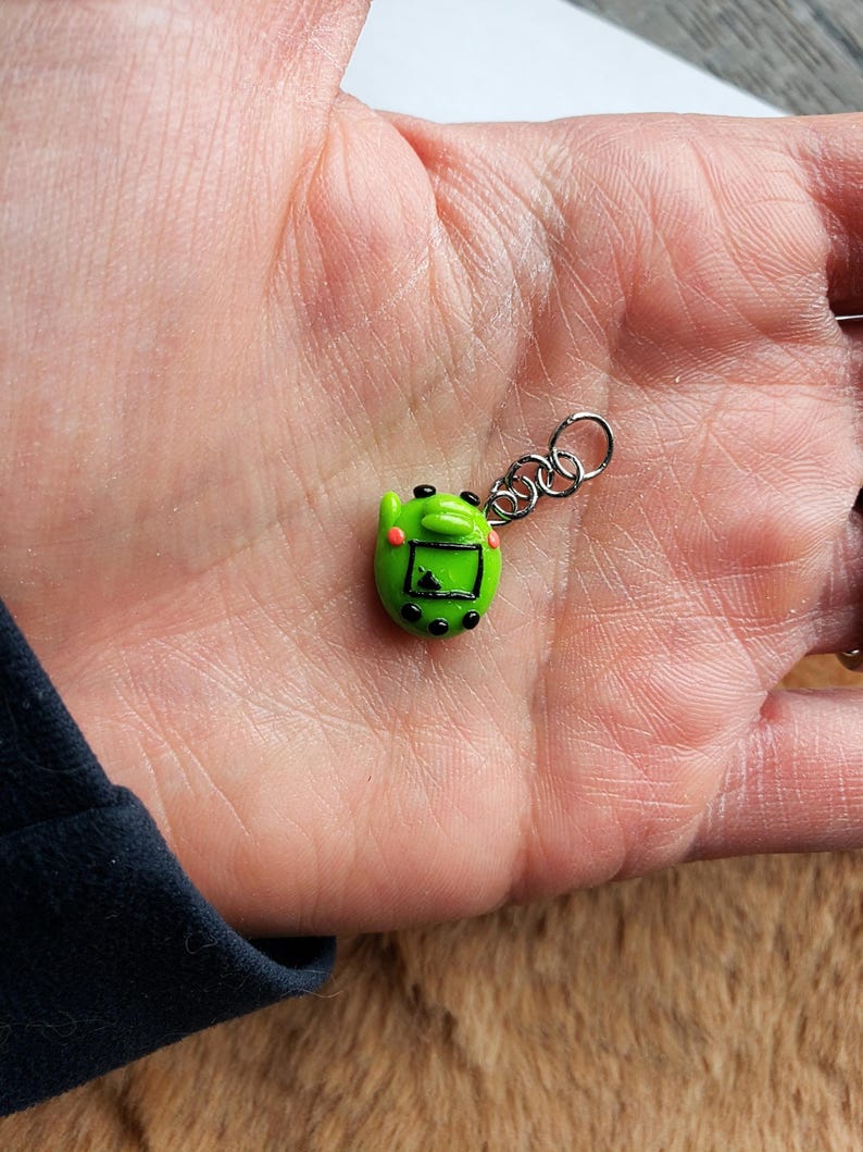May include: A small, green, handheld video game charm with a silver chain. The charm has a screen, black buttons, and pink cheeks. The charm is a miniature version of a retro video game.