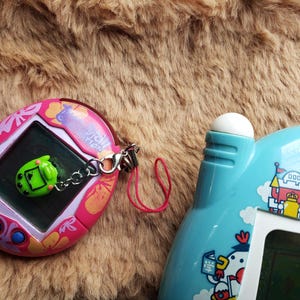 May include: Two vintage handheld electronic games. One is pink with a screen and a green charm. The other is light blue with a white button and a screen. Both are retro toys.
