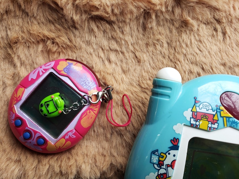 May include: Two vintage handheld electronic games. One is pink with a screen and a green charm. The other is light blue with a white button and a screen. Both are retro toys.