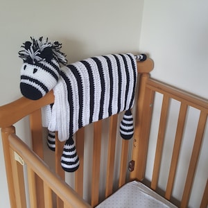 May include: A black and white striped crocheted zebra crib toy hanging from a wooden crib. The zebra has a long tail and a head with black eyes and a black nose.