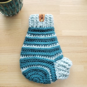 May include: A teal and light blue crocheted dog sweater with a brown leather tag. The sweater has a striped pattern and a ruffled collar.