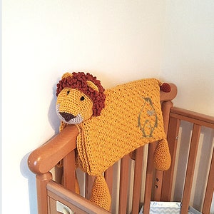 May include: A crocheted lion-shaped baby blanket in yellow and brown with a penguin design on the blanket. The blanket is draped over a wooden cot.
