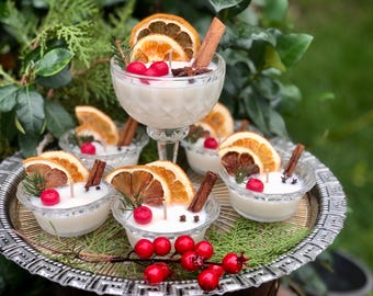 Puede incluir: Un conjunto de cinco velas perfumadas blancas en recipientes de vidrio, decoradas con rodajas de naranja secas, palitos de canela y bayas rojas. Las velas están dispuestas en una bandeja plateada.