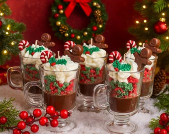 May include: Six clear glass mugs filled with a layered dessert, featuring a dark liquid base, red and green textured middle, and whipped cream topping. Each mug is decorated with a candy cane, gingerbread man, and holly. The scene is set on a snowy surface with Christmas decorations.
