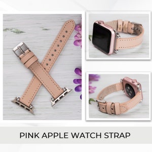 May include: A pink leather Apple Watch strap with silver hardware. The strap is shown on a light wood surface.