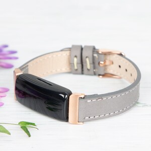 May include: A grey leather band with rose gold hardware for a fitness tracker. The band is stitched with white thread and has a buckle closure.