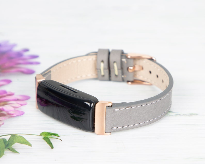 May include: A grey leather band with rose gold hardware for a fitness tracker. The band is stitched with white thread and has a buckle closure.