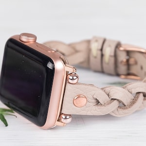 May include: A rose gold Apple Watch with a braided beige leather strap. The strap has a rose gold buckle and decorative accents.