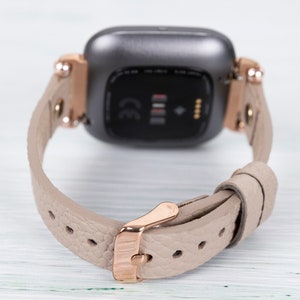 May include: A rose gold smartwatch with a gray face and a beige leather band. The band has a rose gold buckle.