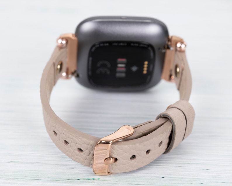 May include: A rose gold smartwatch with a gray face and a beige leather band. The band has a rose gold buckle.