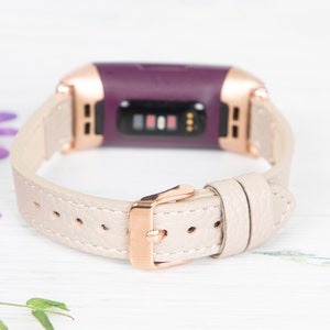 Puede incluir: Una banda de cuero beige claro con una hebilla de oro rosa para un rastreador de fitness morado.