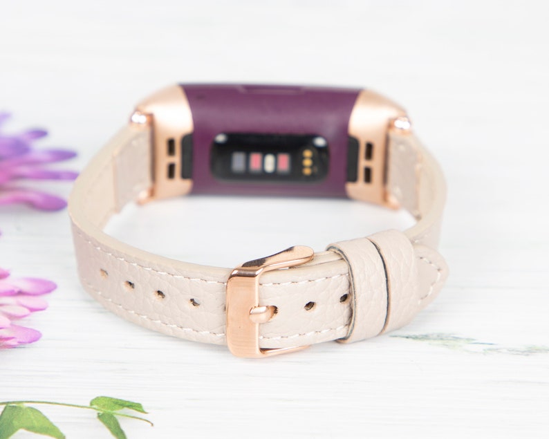 Puede incluir: Una banda de cuero beige claro con una hebilla de oro rosa para un rastreador de fitness morado.