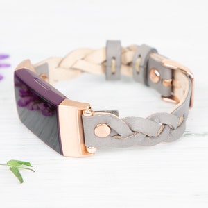 May include: A gray braided leather band with rose gold hardware for a fitness tracker. The tracker is purple and has a black screen.