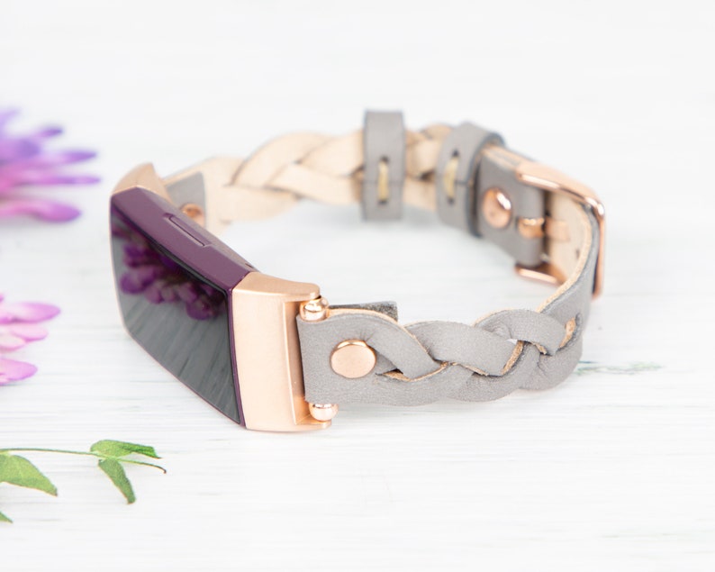 May include: A gray braided leather band with rose gold hardware for a fitness tracker. The tracker is purple and has a black screen.